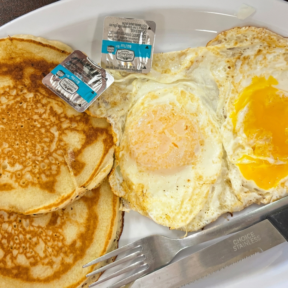 Plate of pancakes and two fried eggs with runny yolks, butter packets, fork, and knife on a restaurant table.
