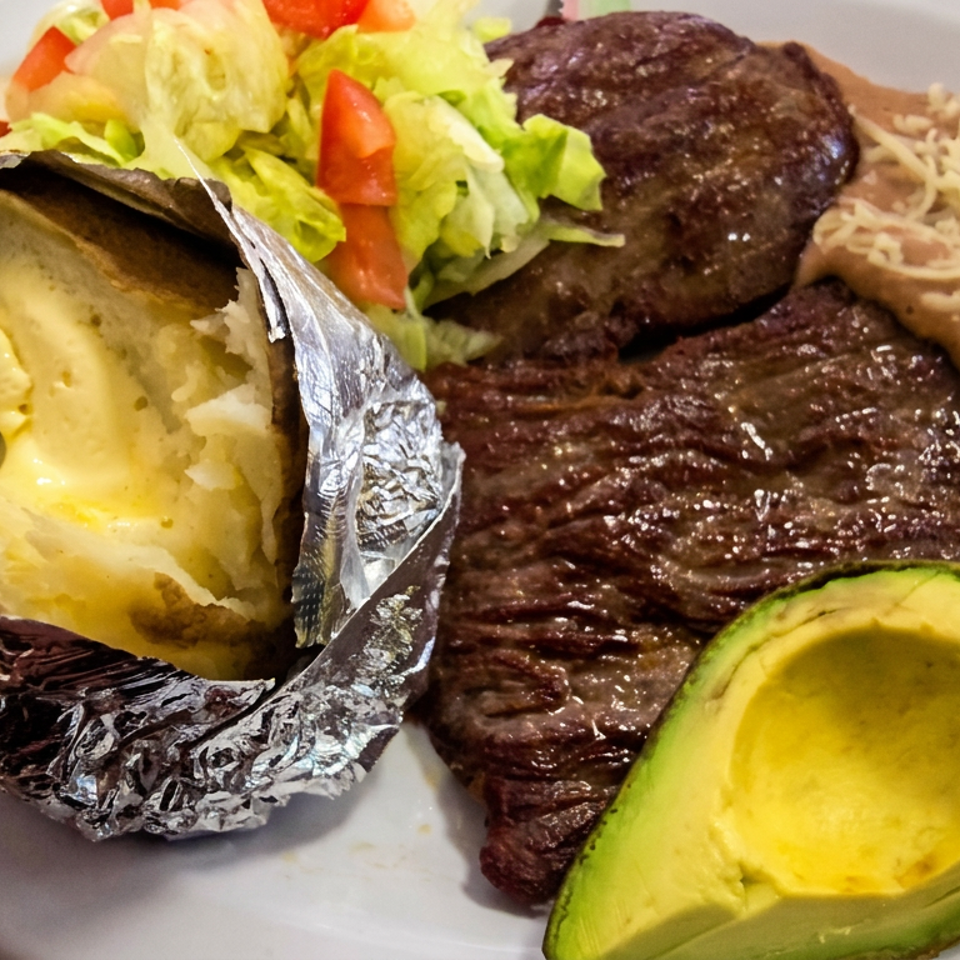 Grilled steak, beef patty, baked potato with butter, rice, salad, and avocado arranged on a white plate.