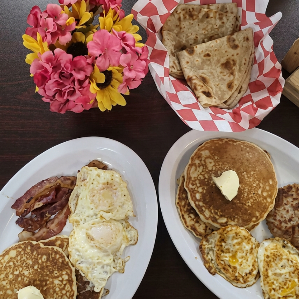 Overhead view of breakfast plates with pancakes, bacon, fried eggs, and savory flatbreads, accompanied by flowers and a wooden serving board.